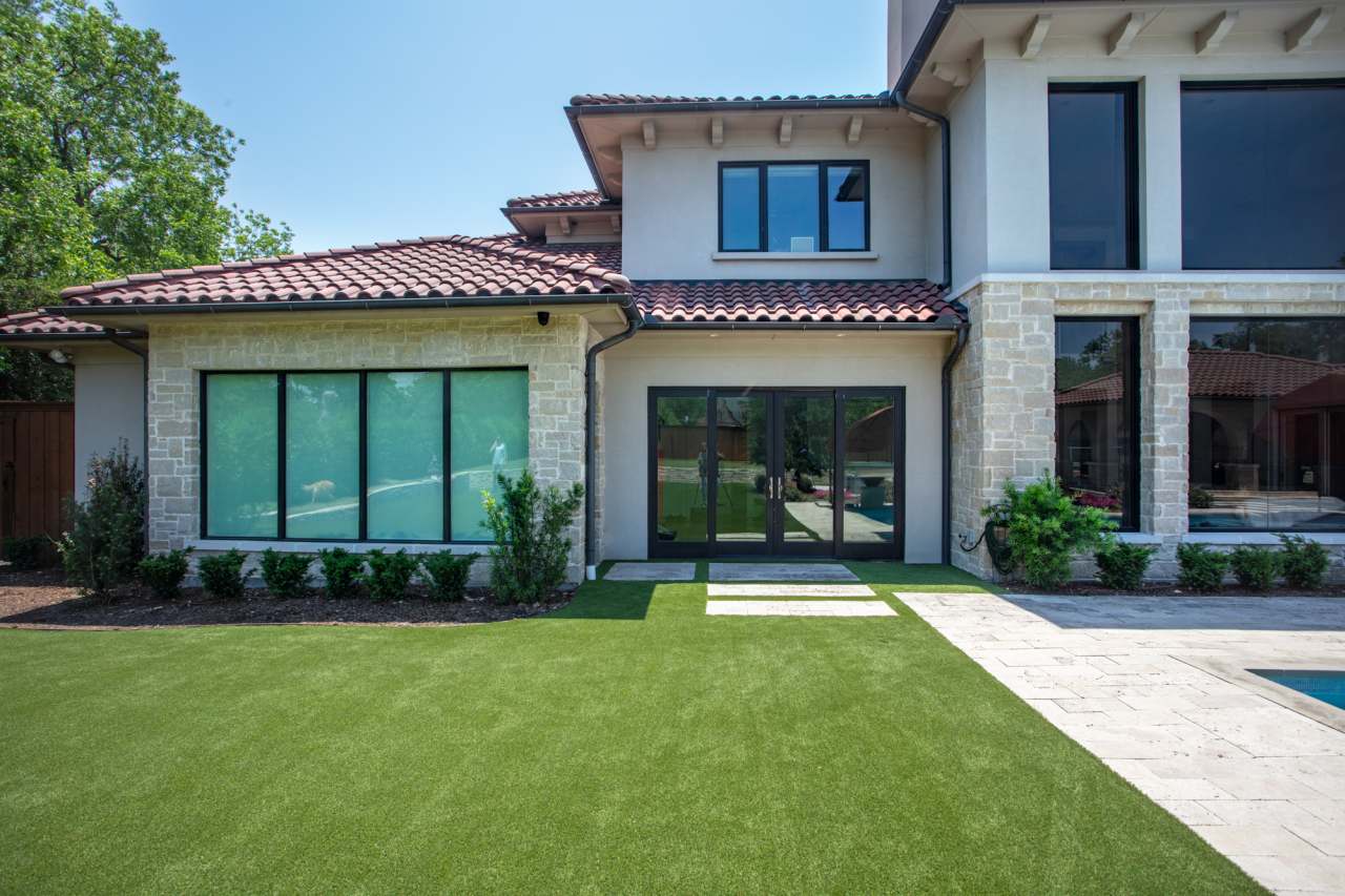 A home’s backyard with artificial turf and stone paths bordered by shrubs and large windows.