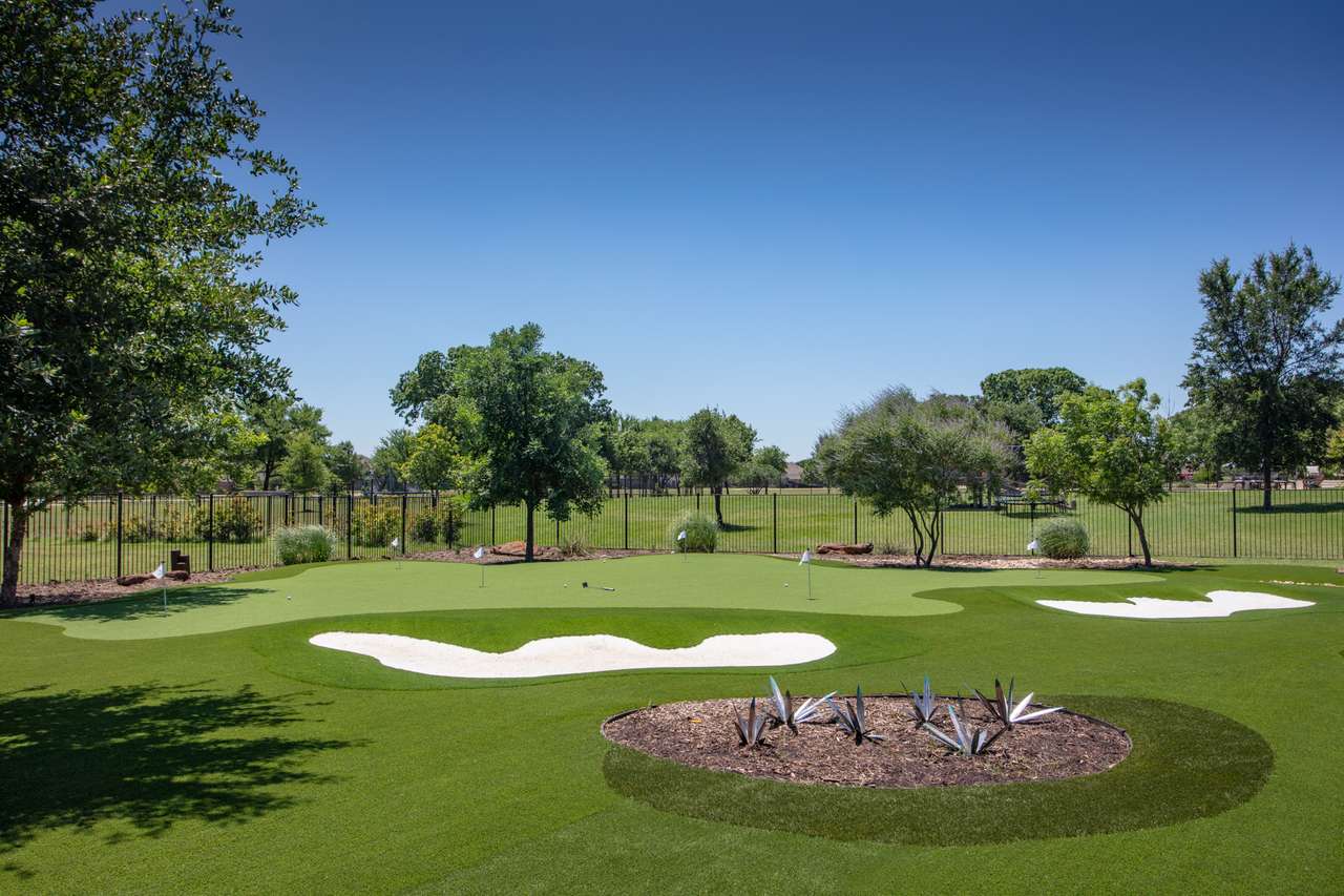Large backyard putting green with multiple holes, sand traps, and turf mounds, surrounded by trees and fenced open parkland.