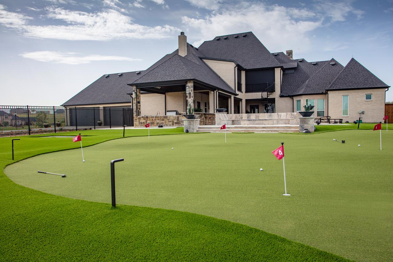 Luxury home with a large, curved backyard putting green featuring red flags, practice golf balls, clubs, and metal ball return ramps.