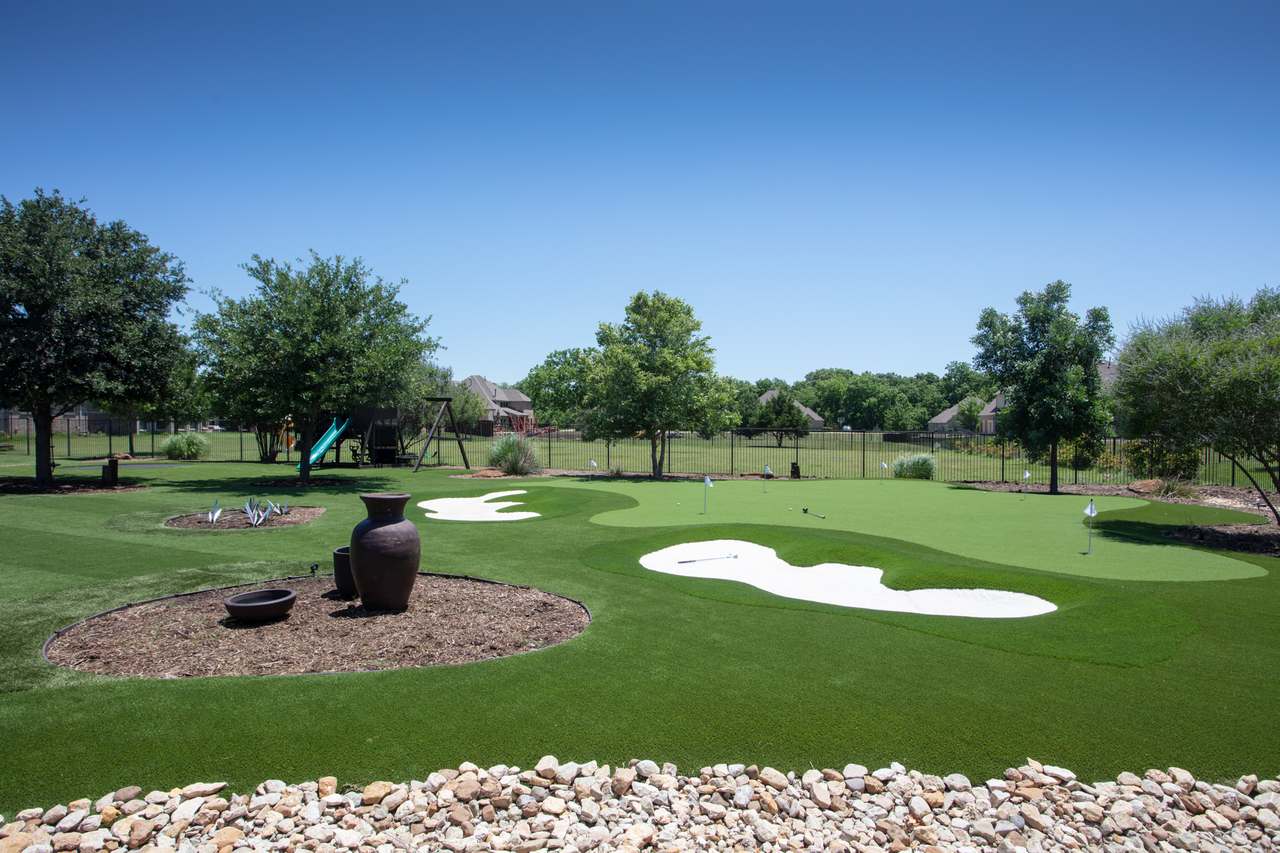 A backyard putting green in Utah with white sand traps, golf flags, pottery, and trees under a bright blue sky.