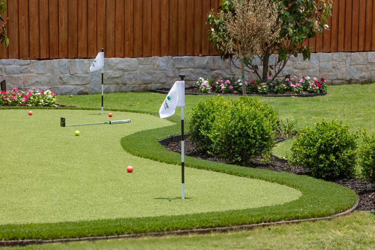 A backyard putting green in Utah with white flags, colorful balls, bushes, flowers, and a wood-and-stone fence.