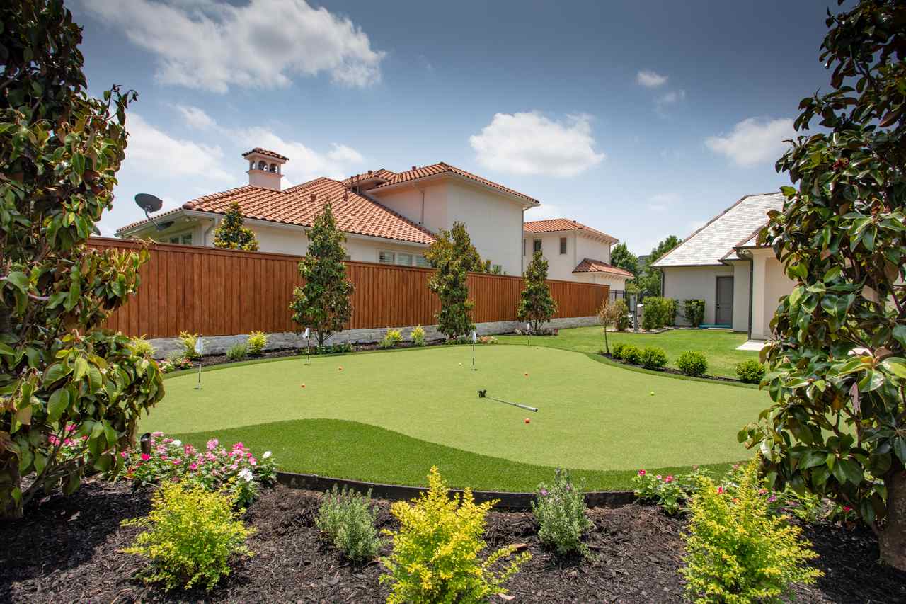 A neatly landscaped backyard features a smooth putting green surrounded by flowers, bushes, and trees, with kids' golf balls and a club scattered on the turf, set against modern homes with red-tiled roofs and a tall wooden fence.