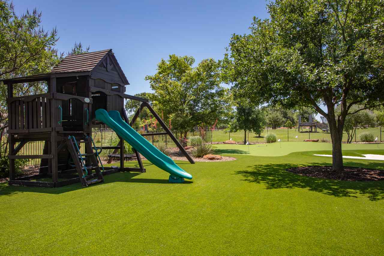 Backyard putting green in Utah with a kids' playset, green slide, trees, and a sand trap on artificial turf.