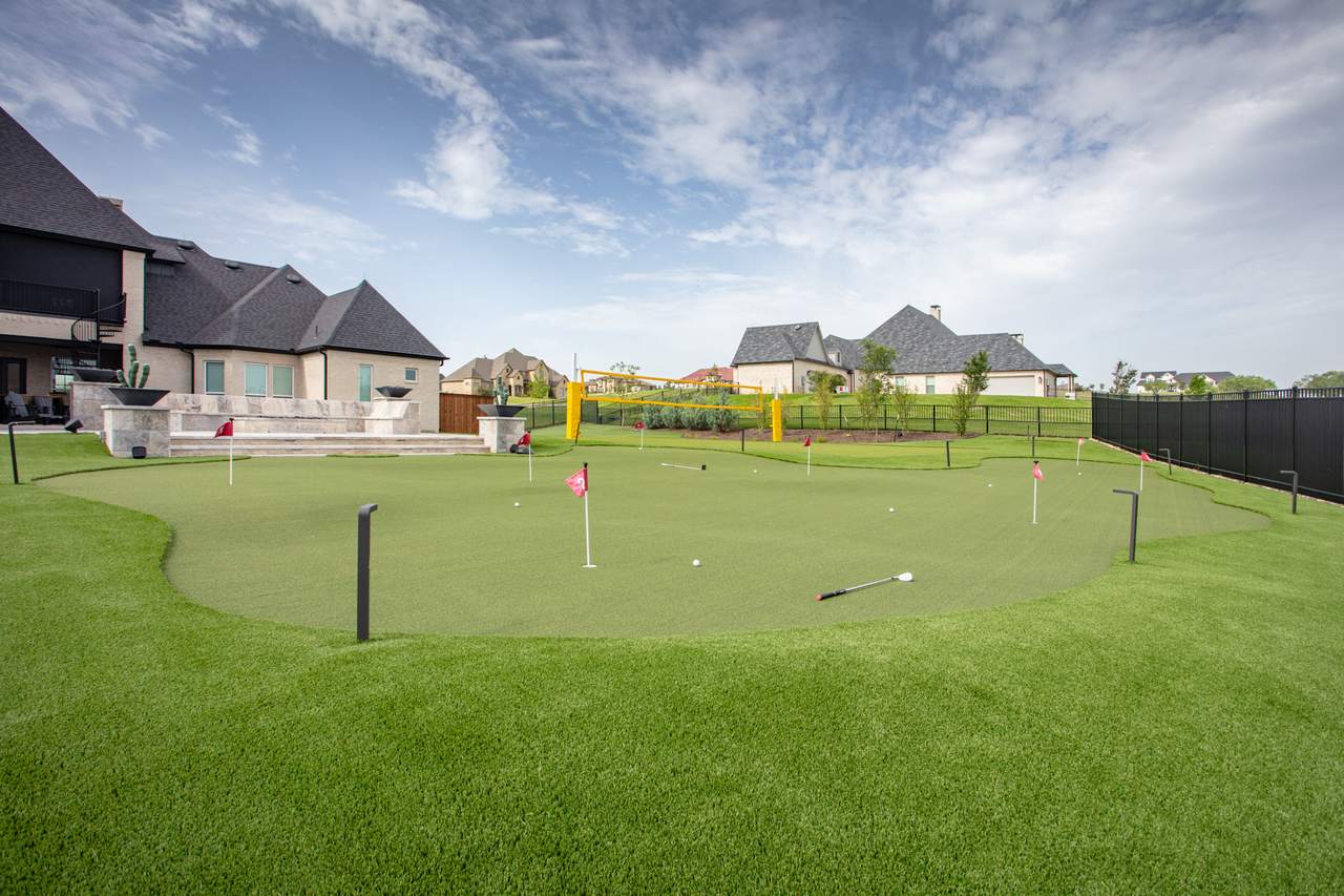 A backyard putting green in Utah with red flags and golf clubs, surrounded by houses and a black metal fence.