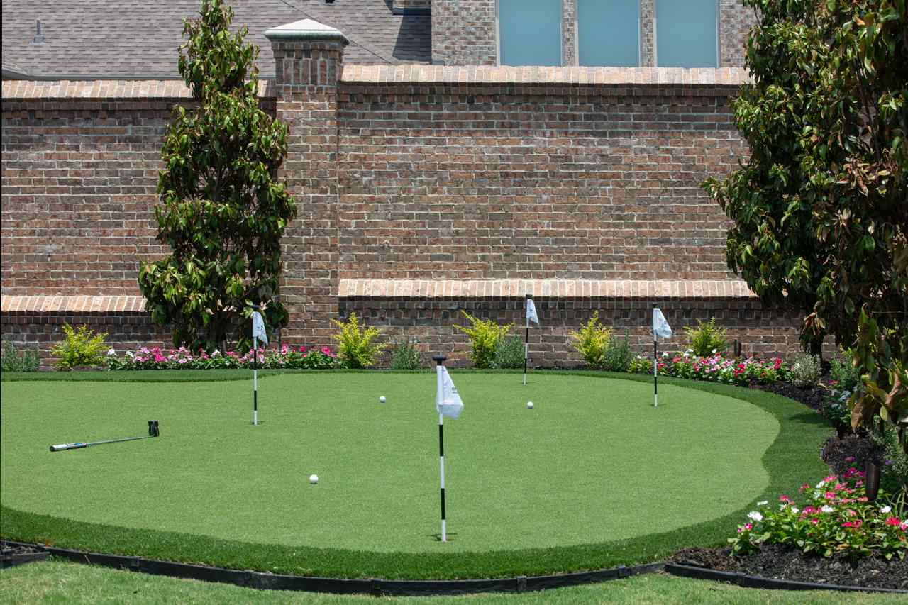A backyard putting green in Utah with white flags and golf balls, bordered by brick walls, flowers, and trees.