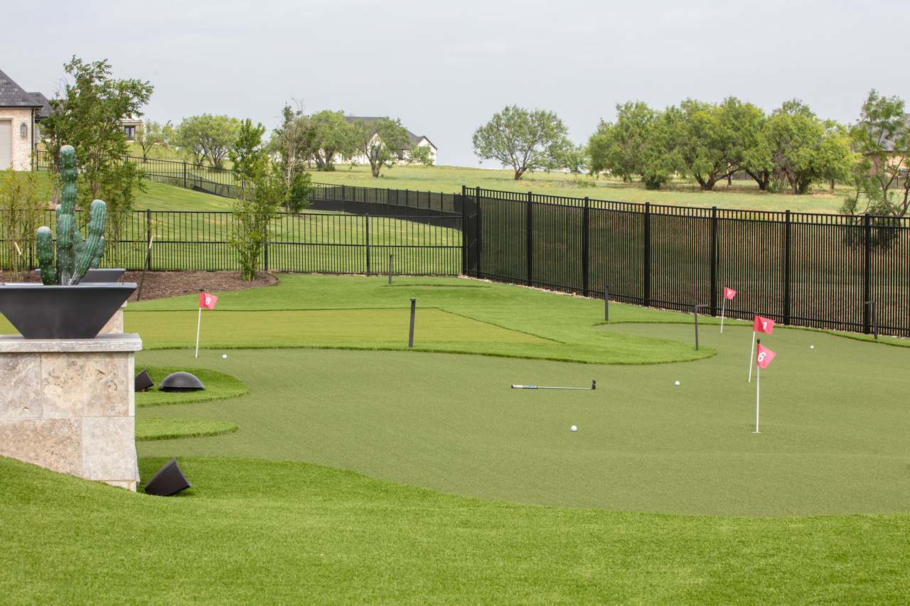 A backyard putting green in Utah with small flags and golf balls, surrounded by a black fence and greenery.